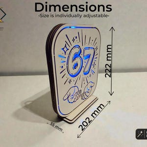 May include: A laser-cut wooden sign featuring the number 67, star accents, and two hands, illuminated by blue light. The sign measures 222 mm in height, 202 mm in width, and 55 mm in depth. Text includes "Dimensions" and "Size is individually adjustable."