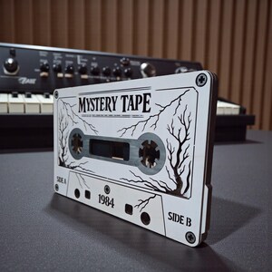 May include: A white cassette tape with the text "MYSTERY TAPE" and "1984" printed on it. The cassette tape has a black and white tree design. The background includes a black synthesizer.