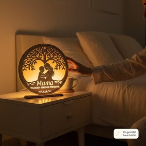 Puede incluir: Una l&aacute;mpara circular de madera con un dise&ntilde;o cortado a l&aacute;ser de un &aacute;rbol con la silueta de un progenitor y un ni&ntilde;o debajo. La l&aacute;mpara emite una luz c&aacute;lida y muestra el texto Mama Wurzel meines Herzens en un acabado de madera clara.