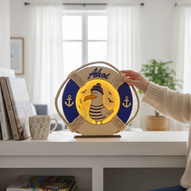 Pu&ograve; includere: Lampada decorativa a forma di salvagente, con un motivo a gabbiano dei cartoni animati con cappello e camicia a righe. La parola "Ahoi" &egrave; scritta sopra il gabbiano. La lampada ha una combinazione di colori blu e crema con dettagli di corda e ancore.