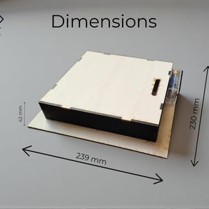 May include: A wooden box with a black base and a light-colored top. The dimensions are 9.4 inches long, 9 inches wide, and 1.7 inches high. The box has a slot on the top and a clear plastic tube attached to the side.