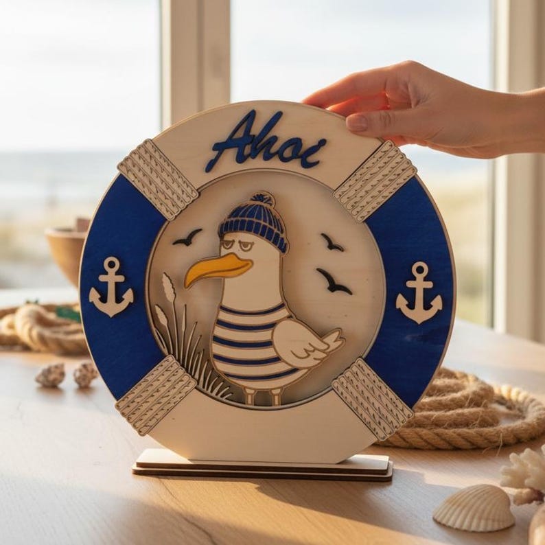 Pu&ograve; includere: Decorazione nautica in legno con un gabbiano con una camicia a righe blu e bianca e un berretto blu. Il gabbiano &egrave; all'interno di un salvagente blu e bianco con la scritta "Ahoi" e dettagli di ancora.