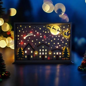 May include: A white wooden Christmas light-up scene with a silhouette of Santa Claus and reindeer flying over a moon with stars and a small town with houses and streetlights.