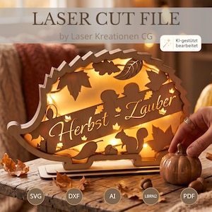 Op de afbeelding: Een lasergesneden houten decoratie in de vorm van een egel met de tekst "Herbst - Zauber" en uitgesneden bladeren, een eekhoorn en een eikel. Het object wordt van binnenuit verlicht door warme lichtjes en staat op een houten voet.