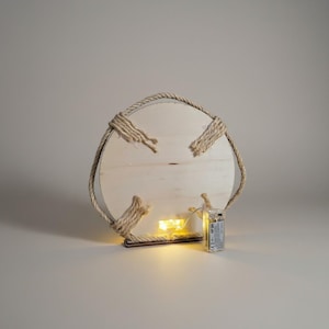 Pu&ograve; includere: Una decorazione da parete rotonda in legno con finitura legno naturale. &Egrave; circondata da una spessa corda e presenta quattro accenti di corda. Piccole luci LED bianche calde sono attaccate sul retro, alimentate da un pacco batterie.