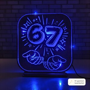May include: A rectangular, dark-colored light box featuring a glowing blue LED design. The center displays the number 67 surrounded by radiating lines and star shapes, with two open hands illustrated below the number, all illuminated in bright blue.