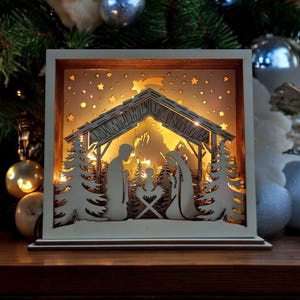 Könnte beinhalten: Eine weiße Holz-Krippe mit einem Licht im Inneren. Die Szene zeigt Maria, Josef und das Jesuskind in einer Krippe. Die Szene wird von einem warmweißen Licht beleuchtet.