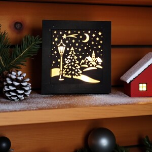 May include: A black, square, illuminated Christmas scene with a cutout design of a streetlight, trees, houses, moon, and stars. A small red house with a white snow-covered roof sits to the right. Pinecones and greenery are on the left.