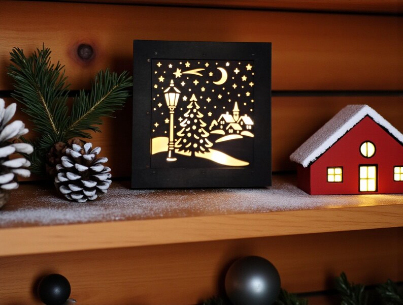 May include: A black, square, illuminated Christmas scene with a cutout design of a streetlight, trees, houses, moon, and stars. A small red house with a white snow-covered roof sits to the right. Pinecones and greenery are on the left.