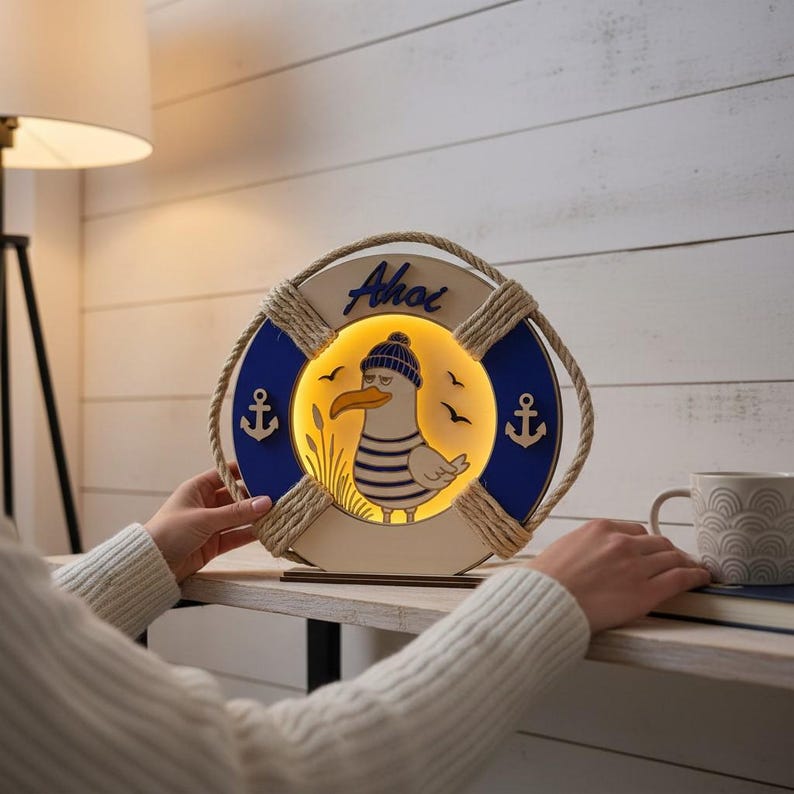 Pu&ograve; includere: Lampada decorativa in legno a forma di salvagente, con un design a forma di gabbiano dei cartoni animati che indossa un cappello e una camicia a righe. Il design include la parola "Ahoi", sezioni blu e bianche, dettagli di corda e simboli di ancora.