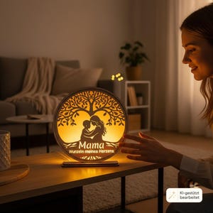Puede incluir: Una l&aacute;mpara circular de madera con un dise&ntilde;o cortado a l&aacute;ser de un &aacute;rbol con la silueta de un padre o madre y un ni&ntilde;o en la base. La l&aacute;mpara emite una luz c&aacute;lida y muestra el texto "Mama" y "Wurzeln meines Herzens" en letra cursiva.