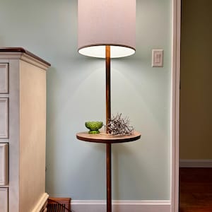 May include: A mid-century modern floor lamp with a round lampshade and a small side table. The lamp has a wooden base and a brass accent. A green glass bowl and dried plant sprigs sit on the table.
