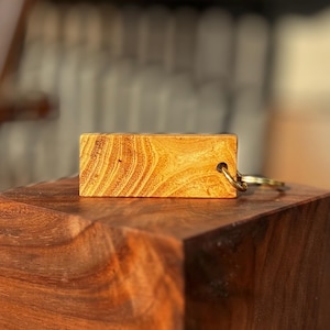 Może przedstawiać: Prostokątny drewniany brelok do kluczy z naturalnym wzorem słojów drewna. Brelok ma jasnobrązowy kolor i ma złoty metalowy pierścień. Brelok spoczywa na ciemnobrązowym drewnianym bloku.