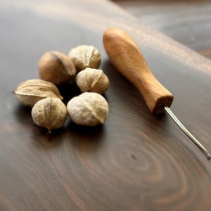 May include: A wooden nut pick with a metal tip rests beside a small pile of nuts on a dark wooden surface. The nuts are light brown and have textured shells. The nut pick has a smooth, light-colored wooden handle.
