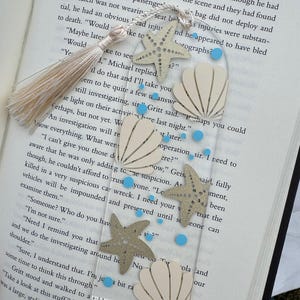 May include: Clear acrylic bookmark with a beige tassel, starfish, and seashell cutouts, and small blue dots. The bookmark is placed on an open book, adding a touch of coastal charm to reading.