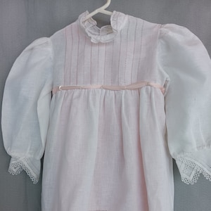 May include: White linen dress with a pink ribbon and lace trim. The dress has a ruffled collar and long sleeves.