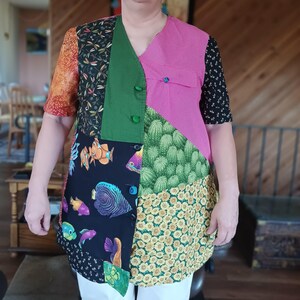 May include: A patchwork tunic with a V-neck design. The tunic features a variety of fabric patterns, including fish, floral, and geometric prints. The color palette includes pink, green, black, orange, and yellow. The tunic has short sleeves and is buttoned down the front.