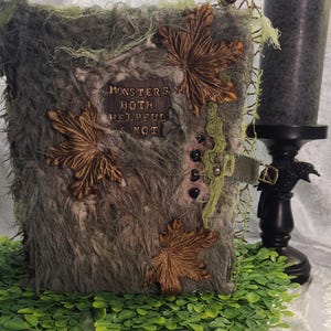 May include: A rectangular book with a textured, shaggy gray cover decorated with three brown fabric leaves, green moss-like fibers, and a central plaque reading "MONSTERS BOTH HELPFUL & NOT". The book features a side strap with a metal buckle.