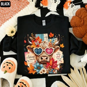 May include: A black long-sleeve sweatshirt featuring a graphic of a Highland cow wearing heart-shaped glasses. The design includes colorful autumn leaves, flowers, and the text "Autumn leaves" and "the leaves fall one by one."