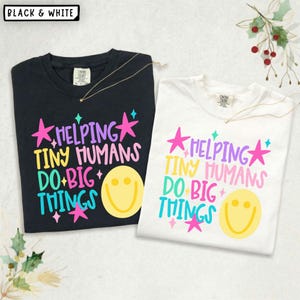 May include: A black t-shirt and a white t-shirt, each featuring the colorful text "HELPING TINY HUMANS DO BIG THINGS" in purple, yellow, pink, and teal, accompanied by pink and teal star icons and a large yellow smiley face graphic.