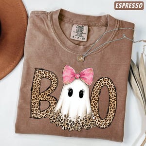 May include: An espresso-colored t-shirt featuring a graphic of a white ghost with a pink bow and the word BOO in a leopard print pattern. The shirt has a crew neckline and a Comfort Colors label at the inner collar. A silver chain necklace is visible.