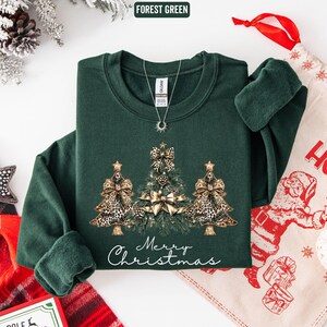 May include: A forest green crewneck sweatshirt featuring a graphic of three Christmas trees decorated with gold leopard print bows and pinecones. The text Merry Christmas is printed in white script below the trees. A silver necklace rests on the collar.