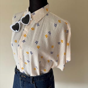May include: A short-sleeved button-down shirt in cream with a pattern of small blue and yellow flowers and tiny brown dots, paired with a dark blue denim skirt. A pair of white-framed heart-shaped sunglasses is attached to the shirt collar.
