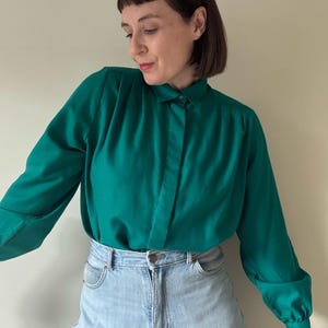 May include: A long-sleeved emerald green button-down shirt featuring a pointed collar, a concealed front placket, and gathered shoulder detailing. The shirt is paired with light-wash blue denim jeans that have a high-waisted fit and metal hardware.