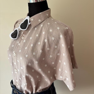 May include: A short-sleeved, light brown button-down shirt featuring a white polka dot pattern, paired with dark blue denim jeans. A pair of white-framed heart-shaped sunglasses with dark lenses is resting on the collar of the shirt.