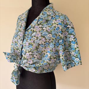 May include: A short-sleeved button-down shirt featuring a vibrant floral pattern in shades of blue, light purple, and white against a lime green background. The shirt has a classic collar and is tied at the front waist with a knot detail.