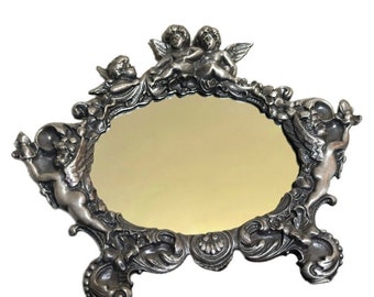 May include: A silver-toned ornate mirror with a cherub design. The mirror is oval shaped and has a decorative frame with cherubs and floral details.
