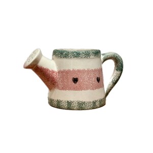 May include: A ceramic watering can with a white base, pink stripe, and green speckled accents. The watering can features a spout, handle, and two black heart designs. A charming garden decor item.