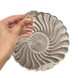 May include: A clear glass dish with a swirl design. The dish is shaped like a seashell and has a scalloped edge.