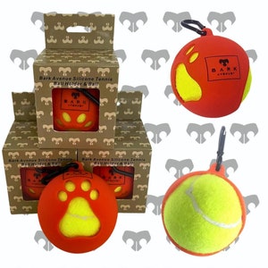 以下が含まれることがあります： 黄色いテニスボールが入った赤いシリコン製のテニスボールホルダーが3つ。ホルダーには肉球のデザインと「Bark Avenue」の文字が書かれています。