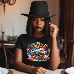 May include: Black t-shirt featuring a colorful graphic of a train with the text "Soul Train". The design includes a rainbow-colored platform and disco ball elements. The person in the image is wearing a black hat.