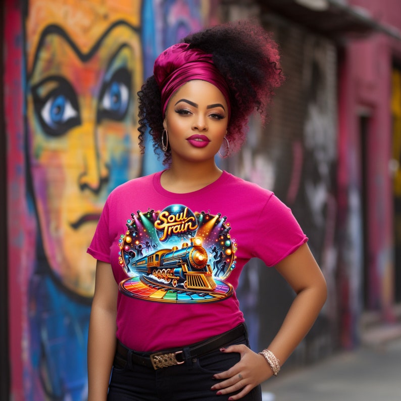 May include: A magenta t-shirt with a colorful graphic of a train, stage lights, and the words "Soul Train". The design is set against a rainbow-colored track. The t-shirt is worn by a person in front of a colorful mural.