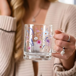 May include: Clear glass mug with a handle, adorned with a floral design featuring pink, purple, and white flowers. The mug is held by a person wearing a beige sweater. The mug is perfect for enjoying your favorite beverage.