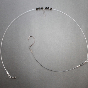 May include: Fishing rig with a clear monofilament line, a fishing hook, and a black wire leader. The rig includes a swivel and a series of black beads. The components are arranged against a gray background.