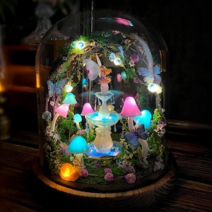 Peut inclure: Une sc&egrave;ne f&eacute;erique miniature sous un d&ocirc;me en verre. Le paysage comprend une fontaine, des champignons color&eacute;s, des papillons et une figurine de f&eacute;e. L'ensemble est &eacute;clair&eacute; par des lumi&egrave;res LED, cr&eacute;ant une ambiance magique. Le d&ocirc;me repose sur une base en bois.