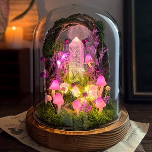 May include: A glass dome cloche on a round wooden base containing a miniature forest scene with a central clear crystal point, green moss, twisted branches, and several small, glowing pink mushroom-shaped lights among purple floral accents.