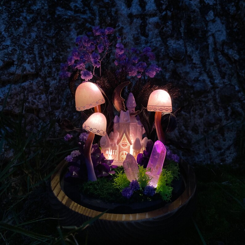 May include: A miniature fairy house with a lit window, surrounded by purple flowers, crystals, and glowing mushrooms in a wooden planter.