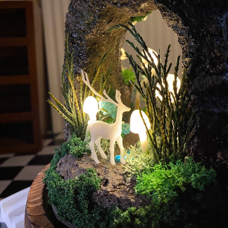 May include: A white ceramic deer figurine standing in a miniature forest scene. The scene is made of moss, small trees, and white glowing mushrooms. The deer is standing on a rock base.