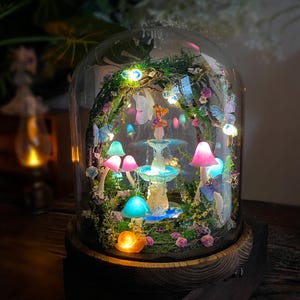 Peut inclure: Une sc&egrave;ne de jardin f&eacute;erique miniature enferm&eacute;e dans un d&ocirc;me en verre. Le paysage miniature comprend une fontaine, des champignons color&eacute;s, des papillons et une figurine de f&eacute;e, le tout &eacute;clair&eacute; par de petites lumi&egrave;res. La base est en bois fonc&eacute;.