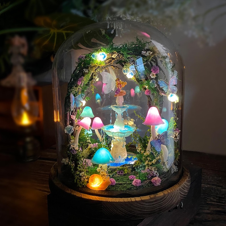 Peut inclure: Une sc&egrave;ne de jardin f&eacute;erique miniature enferm&eacute;e dans un d&ocirc;me en verre. Le paysage miniature comprend une fontaine, des champignons color&eacute;s, des papillons et une figurine de f&eacute;e, le tout &eacute;clair&eacute; par de petites lumi&egrave;res. La base est en bois fonc&eacute;.