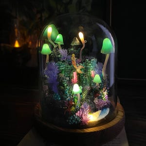 May include: A decorative glass dome terrarium with a miniature underwater scene, including a castle, a mermaid, and glowing green mushrooms. The wooden base supports the glass dome. The scene is lit with colourful LED lights, creating a magical atmosphere.