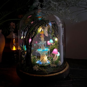 Peut inclure: Une sc&egrave;ne de jardin f&eacute;&eacute;rique miniature sous un d&ocirc;me en verre. Le paysage comprend une fontaine lumineuse, des champignons color&eacute;s et des guirlandes lumineuses. La base est un cercle en bois. La sc&egrave;ne est &eacute;clair&eacute;e par des lumi&egrave;res douces et color&eacute;es.
