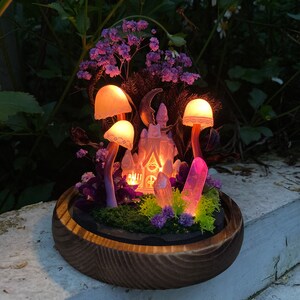May include: A miniature fairy house with a pink and white exterior, surrounded by purple flowers, glowing mushrooms, and crystals, all set in a wooden bowl.