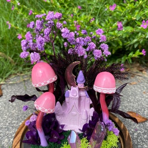 May include: A miniature fairy garden scene with a pink castle, pink mushrooms, purple flowers, and iridescent crystals in a wooden planter.