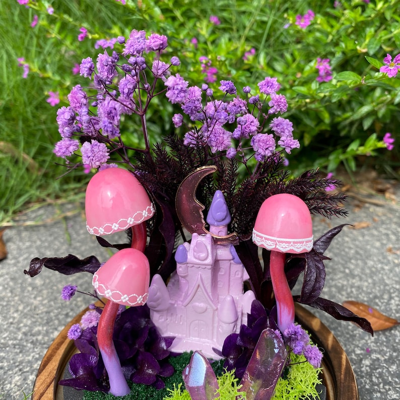 May include: A miniature fairy garden scene with a pink castle, pink mushrooms, purple flowers, and iridescent crystals in a wooden planter.