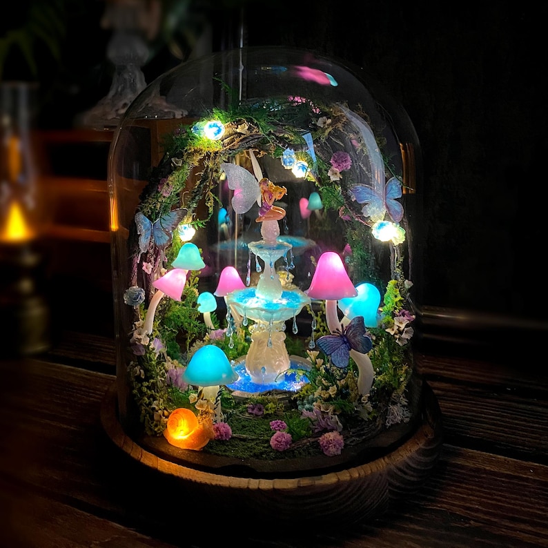 Peut inclure: Une sc&egrave;ne f&eacute;erique miniature sous un d&ocirc;me en verre. Le paysage comprend une fontaine, des champignons color&eacute;s, des papillons et une figurine de f&eacute;e. L'ensemble est &eacute;clair&eacute; par des lumi&egrave;res LED, cr&eacute;ant une ambiance magique. Le d&ocirc;me repose sur une base en bois.
